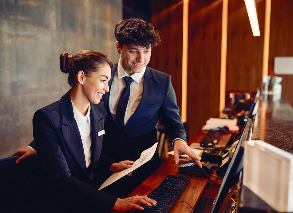 Två hotellreceptionister som samarbetar vid en dator, vilket representerar teamwork, effektiv schemaläggning och arbetsstyrning